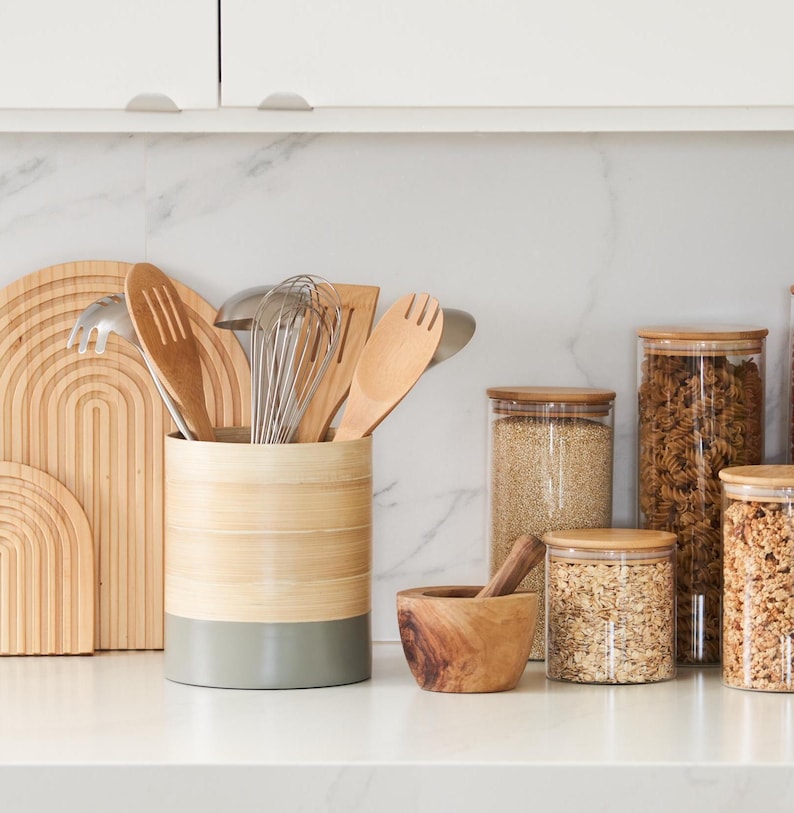 May include: A bamboo utensil holder with a gray base, filled with wooden spoons, a fork, a ladle, and a whisk. The holder is on a white countertop next to a wooden mortar and pestle and four glass jars with lids, filled with various grains and cereals.