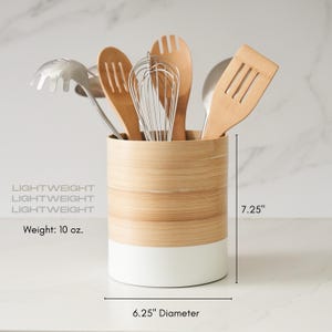 May include: A white and natural bamboo utensil holder with a white base and a natural bamboo top. The holder is 7.25 inches tall and 6.25 inches in diameter. It is holding a variety of kitchen utensils, including a whisk, a wooden spoon, a spatula, and a slotted spoon. The holder weighs 10 ounces.