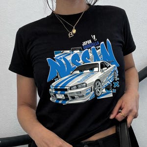 Könnte beinhalten: Schwarzes T-Shirt mit einer Cartoon-Illustration eines silbernen und blauen Nissan Skyline GT-R. Das Shirt zeigt das Wort "NISSAN" in Blau und den Text "JAPAN GOODZELLA".