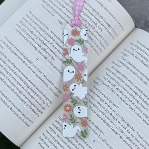 May include: A pink and white gingham bookmark with a clear plastic charm featuring a floral design and multiple white ghosts. The bookmark is placed on an open book with text about faeries and pigs.