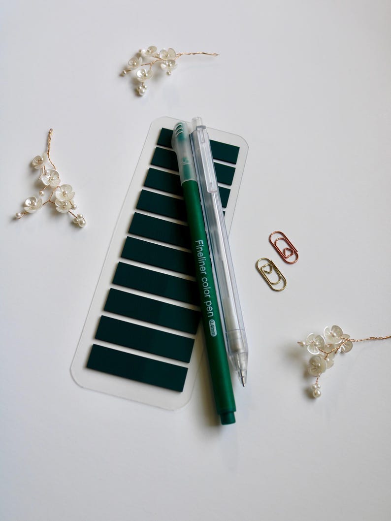 Dark green annoation tab sheet with one transparent mechanical pencil, one blue felt tip pen and two mini paper clip (gold and rose gold) on a white surface. This elementas are part of the mini kit offering. Annotation tab pallet is forest veil.