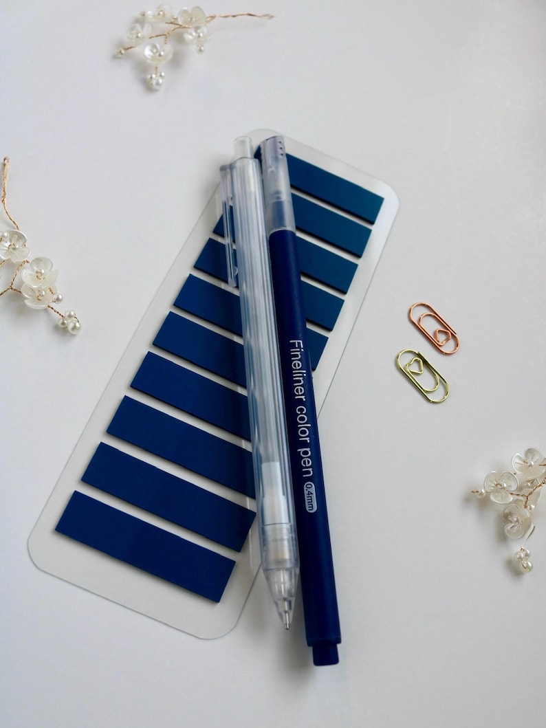 Dark blue annoation tab sheet with one transparent mechanical pencil, one blue felt tip pen and two mini paper clip (gold and rose gold) on a white surface. This elementas are part of the mini kit offering. Annotation tab pallet is Dreamy Lagoon.