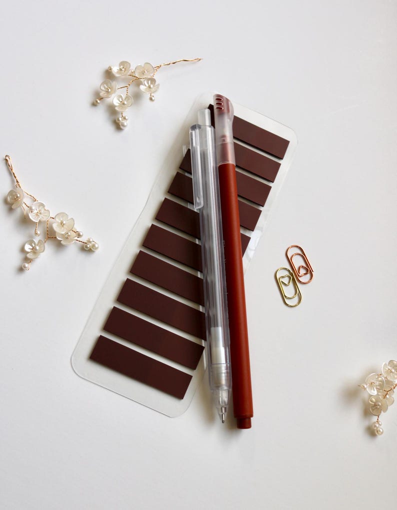 Brown annoation tab sheet with one transparent mechanical pencil, one blue felt tip pen and two mini paper clip (gold and rose gold) on a white surface. This elementas are part of the mini kit offering. Annotation tab pallet is mystic oak.