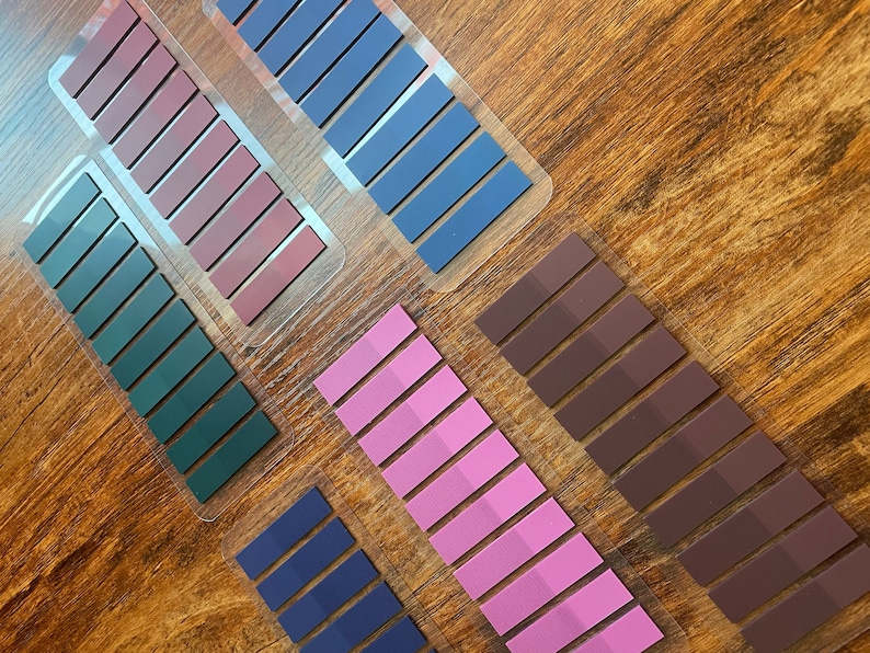 Assortment of colorful annotation tab sheets, each sheet features multiple rectangular strips in shades of pink, purple, dark brown, dark green, and navy blue.  The tapes are neatly arranged on a wooden surface, showcasing their color variations and uniform size. These decorative tapes are ideal for journaling, book annotation, studying and office.