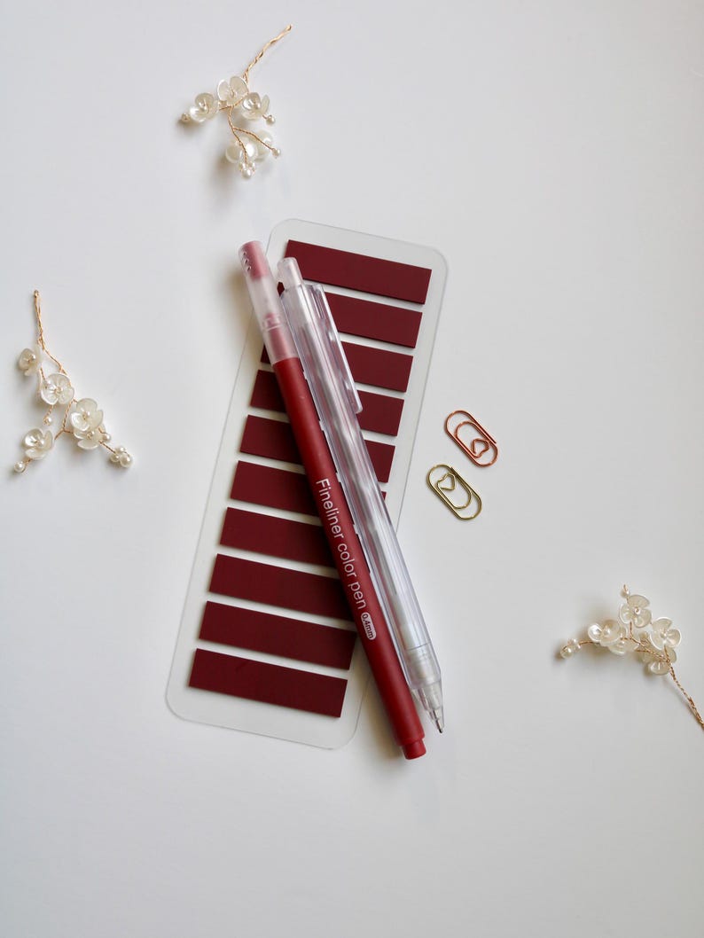 Deep red annoation tab sheet with one transparent mechanical pencil, one blue felt tip pen and two mini paper clip (gold and rose gold) on a white surface. This elementas are part of the mini kit offering. Annotation tab pallet is Blood Moon.