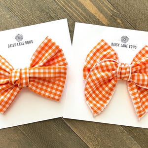 May include: Two orange and white gingham hair bows with white stitching. The bows are on white cards with the text "Daisy Lane Bows".