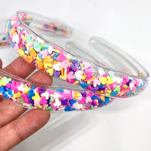 May include: Two clear plastic headbands with colorful sprinkles embedded inside. The sprinkles are in various shapes, including hearts, stars, and dots. The headbands are a popular accessory for girls and women.