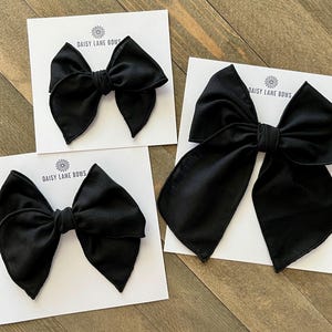 May include: Three black fabric hair bows of different sizes on white cards with the text "Daisy Lane Bows" printed on each card.