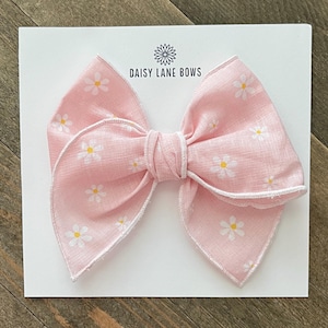 May include: A pink fabric bow with a daisy pattern and white trim, set on a white card. The bow is tied in the center with a matching fabric. The card has the text "DAISY LANE BOWS".