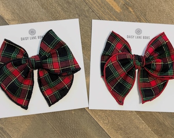 May include: Two large plaid fabric hair bows in red, green, black, and gold, displayed on white cards. The bows have a classic tartan pattern. The cards have the text "DAISY LANE BOWS" printed on them.