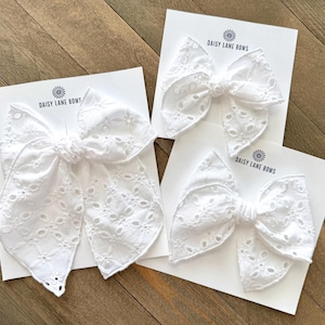 May include: Three white eyelet fabric hair bows on white cards with the text "DAISY LANE BOWS". The bows are arranged on a wooden surface. The bows have a detailed floral pattern.