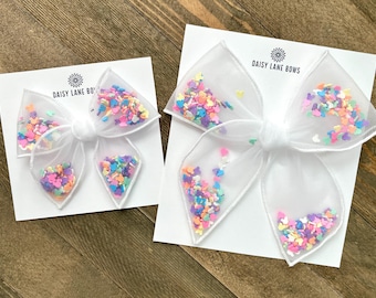 Puede incluir: Dos lazos blancos para el pelo con una superposición transparente llena de confeti colorido en forma de corazón. Los lazos están sujetos a tarjetas blancas con el texto "DAISY LANE BOWS" impreso en negro.