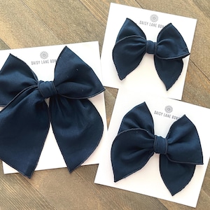 May include: Three navy blue fabric hair bows on white cards, each with the text "DAISY LANE BOWS". The bows are in different sizes and have a classic bow shape. The background is a light brown wooden surface.