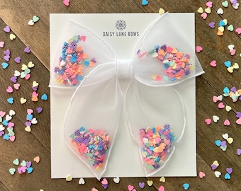 May include: A white tulle hair bow filled with colorful heart-shaped confetti. The bow is centered on a white card with the text "DAISY LANE BOWS". Scattered heart confetti surrounds the bow on a wooden surface.