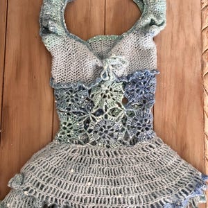 May include: A handmade, light blue crochet top with a unique design. The top features a ruffled skirt, a detailed bodice with floral patterns, and decorative sleeves. The crochet work is intricate, with a mix of light and dark blue yarns.