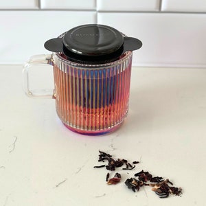 May include: A clear glass tea mug with a black lid and a built-in infuser. The mug has a ribbed design and is filled with a pink-hued tea. Loose tea leaves are scattered on a white countertop.