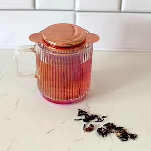 May include: A clear glass mug with a copper-coloured lid and a ribbed design. The mug is filled with a reddish-brown liquid and loose tea leaves are scattered on a white surface.
