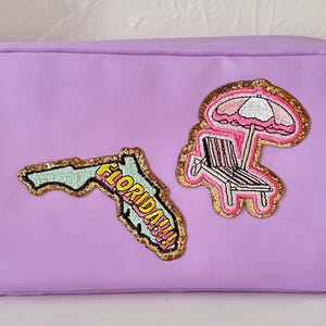 May include: A lilac cosmetic bag with a gold zip. It has two embroidered patches: a map of Florida with the text "FLORIDA!!!" and a beach scene with a chair and parasol.