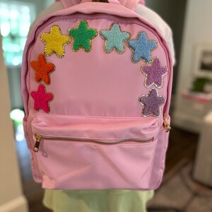 Puede incluir: Una mochila rosa con un bolsillo frontal con cremallera y un arcoíris de estrellas peludas.