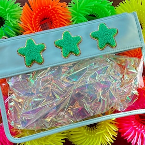 Puede incluir: Una bolsa de cremallera azul claro con un frente de plástico transparente. La bolsa tiene tres adornos en forma de estrella verde en la parte delantera. La bolsa está rodeada de juguetes peludos de colores.