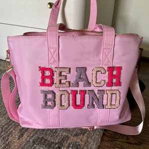 May include: Pink tote bag with a zipper closure and a long strap. The bag has the words "Beach Bound" in colorful, glittery letters.
