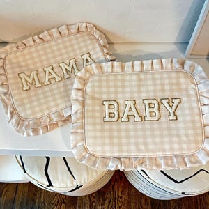May include: Two beige and white gingham pillow covers with ruffled edges. The pillow covers have the words "MAMA" and "BABY" embroidered in gold.