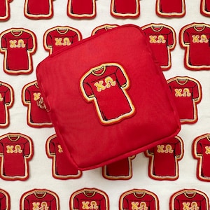 Puede incluir: Bolsa de cosméticos roja con cremallera dorada, con un parche a juego de una camiseta roja con letras doradas. Múltiples parches de camiseta roja con letras doradas rodean la bolsa.