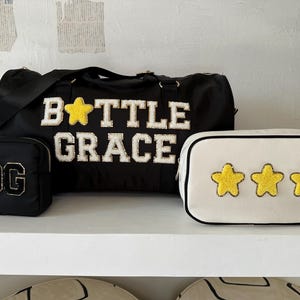 Könnte beinhalten: Drei schwarze und weiße Taschen mit gesticktem Text und gelben Sternen. Die größte Tasche trägt die Aufschrift "Bottle Grace" und einen gelben Stern. Die kleinste Tasche trägt die Aufschrift "OG". Die mittlere Tasche ist weiß mit drei gelben Sternen.