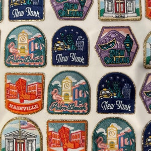 May include: A collection of embroidered patches featuring various cityscapes and landmarks. The patches are colourful and feature a variety of designs, including a pink flamingo, a Statue of Liberty, and a Mardi Gras mask. The patches are all surrounded by a gold glitter border.
