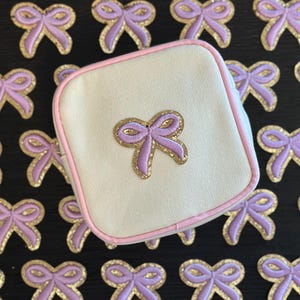 May include: A small, white canvas pouch with a pink trim and a purple bow with gold glitter accents. The pouch is surrounded by many purple bows with gold glitter accents on a black background.