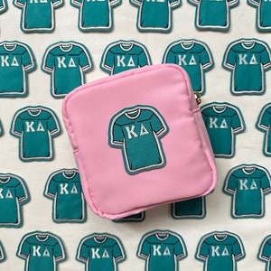 May include: A pink zippered pouch with a teal blue embroidered patch of a t-shirt with the letters "KA" on it. The pouch is surrounded by multiple teal blue embroidered patches of t-shirts with the letters "KA" on them.