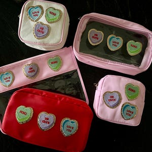 May include: Five pink and white zippered pouches with clear vinyl panels. Each pouch features colorful heart-shaped patches with embroidered text, including "Love You", "Ur Cute", and "XOXO".