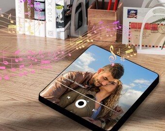 May include: A black square digital photo frame with a couple in a photo on the screen. The photo frame is playing music with the song title "Sweetest Devotion" displayed on the screen. The photo frame is on a wooden surface with musical notes in the background.