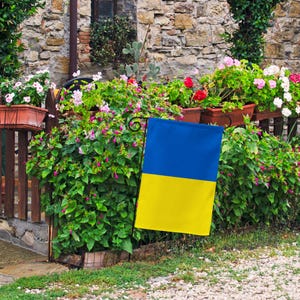 May include: A blue and yellow flag of Ukraine is displayed on a black metal flagpole in front of a stone wall and lush green foliage.