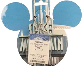 May include: A Space Mountain keychain with a Fastpass ticket. The ticket is in a clear plastic holder and has the text "FASTPASS. Please Return Anytime Between 9:15 AM AND 10:15 AM". The background is a blue sky and the Space Mountain ride.