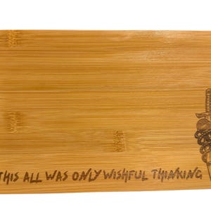 May include: A rectangular bamboo cutting board with rounded corners. The board features an engraved design of a dagger, microphone, and rope, along with the text "THIS ALL WAS ONLY WISHFUL THINKING". The board has a natural wood grain.