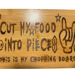 May include: A rectangular bamboo cutting board with rounded corners. The board features engraved text that reads "CUT MY FOOD INTO PIECES" and "THIS IS MY CHOPPING BOARD". Decorative elements include a heart, a star, a skull, a hand gesture, and a symbol.