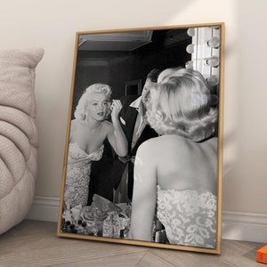 May include: Black and white photograph of a woman applying makeup in front of a mirror. The woman is wearing a strapless dress and has her hair styled in a classic updo. The photograph is framed in a gold frame and is hanging on a white wall.