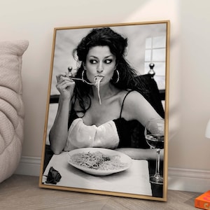 May include: A framed black and white photograph of a woman eating spaghetti. The image is leaning against a wall next to a beige chair and a white lamp. A book with the title "CAPRI DOLCE VITA" is visible.