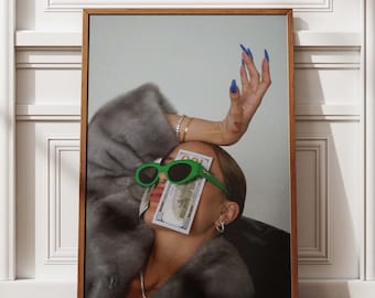 May include: A framed print featuring a person wearing green sunglasses and holding a stack of money over their eyes. The person is wearing a gray fur coat and has blue nail polish. The artwork is displayed against a white wall and a wooden floor.