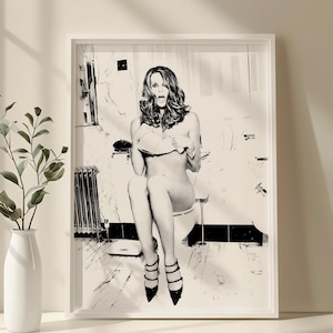 May include: Black and white framed print of a woman sitting on a toilet, with a surprised expression. She is wearing high heels. The artwork is displayed in a white frame, leaning against a wall next to a vase with greenery.