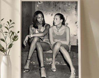 Op de afbeelding: Zwart-witfoto van twee vrouwen die op een trede zitten. De vrouw links draagt een zwart tanktop en de vrouw rechts draagt een rood tanktop. Beide vrouwen dragen kettingen.