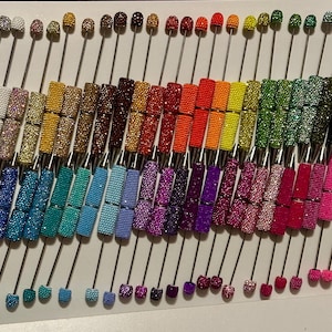 May include: A collection of numerous pens arranged in rows, each featuring a barrel covered in small, sparkling rhinestones in a wide variety of colours including red, blue, green, yellow, pink, purple, black, white, and metallic tones.