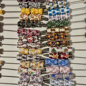 May include: A collection of metal pens featuring decorative barrels adorned with various colourful character prints, patterns, and embedded rhinestones. Each pen has a silver-toned tip and a long, thin metal rod extending from the top end.