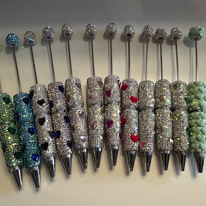 May include: A collection of twelve pens featuring cylindrical barrels encrusted with sparkling rhinestones and inlaid heart-shaped accents in various colours. Each pen is topped with a thin metal rod and a decorative spherical bead topper.