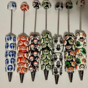 May include: Seven metal pens featuring decorative barrels adorned with various character-themed beads. Each pen has a silver-toned tip and a matching bead topper. The designs include blue, red, black, green, and white patterns with character faces.
