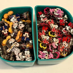 Pode incluir: Dois recipientes de plástico azul-petróleo cheios de pequenos pingentes de acrílico. O recipiente esquerdo contém várias figuras do Mickey Mouse com roupas diferentes, enquanto o direito está cheio de figuras da Minnie Mouse com vestidos.