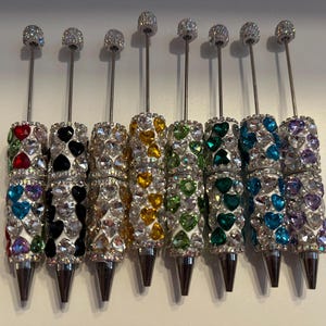 May include: Eight decorative pens with rhinestone-studded barrels in various colours. Each pen has a silver-coloured shaft and tip, and a small, sparkling ball at the top. The barrels are decorated with heart-shaped rhinestones.