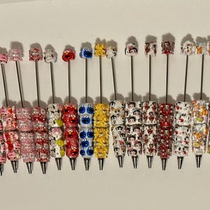 May include: A collection of twenty-one pens arranged in a row, each featuring a silver metal barrel adorned with a stack of colourful, patterned beads. The top of each pen is capped with a small, character-themed topper in various vibrant colours.