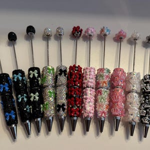 May include: A selection of decorative pens with ornate designs. The pens have silver metal shafts and jewel-covered barrels in colours such as black, red, pink, and white. Some pens feature bow accents, while others have crown or floral toppers.